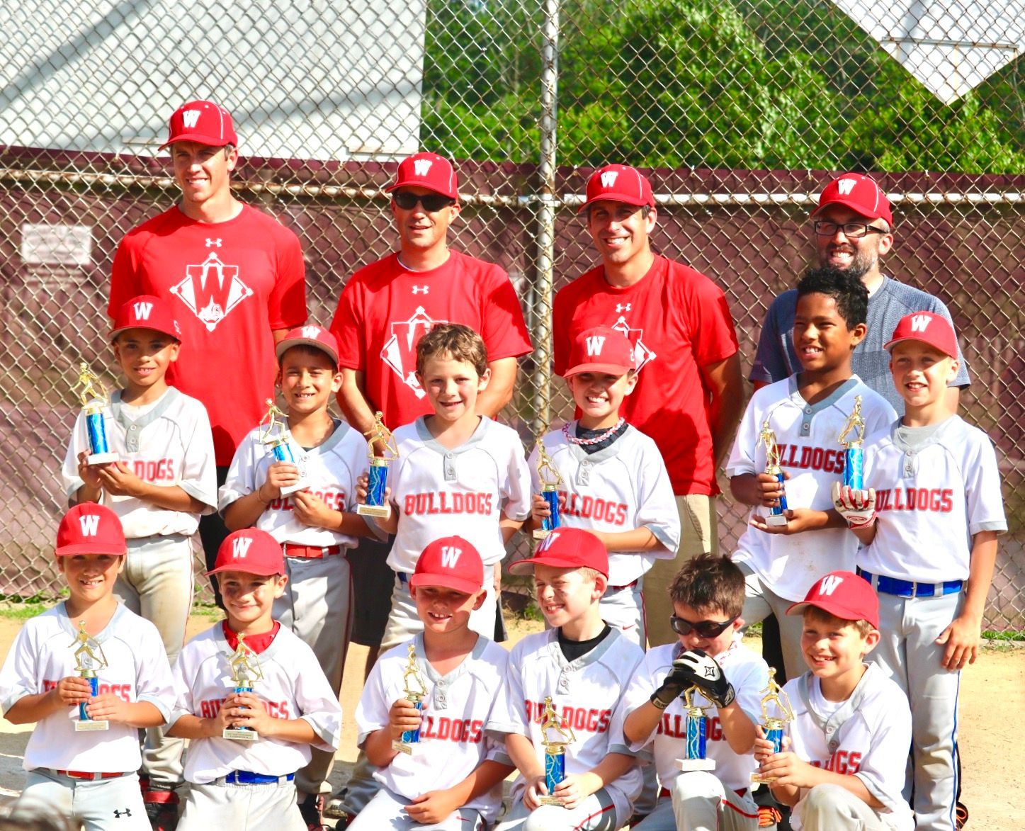 Youth baseball team and coaches posing in red and white uniforms on a field near a chain-link fence.