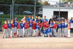 Youth baseball team posing in red and blue uniforms on a dirt field near a fence and dugout