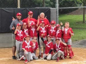 Youth baseball team in red uniforms posing with medals on a dirt field