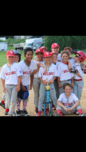 Youth baseball team in white uniforms and red caps posing with a trophy on a dirt field
