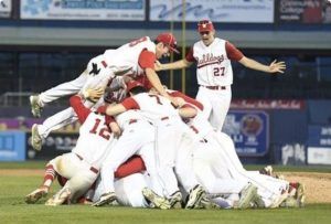 Baseball team in white uniforms celebrating a win in a pile on the field under stadium lights