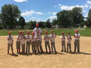 Baseball team posing on a sunny field with a coach, all wearing uniforms and red helmets