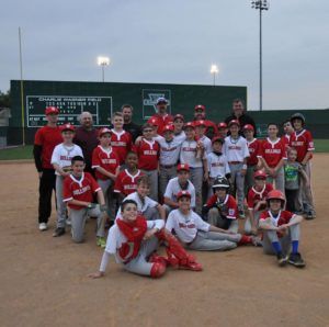 Youth baseball team posing on a dirt field at dusk, wearing red and white uniforms.