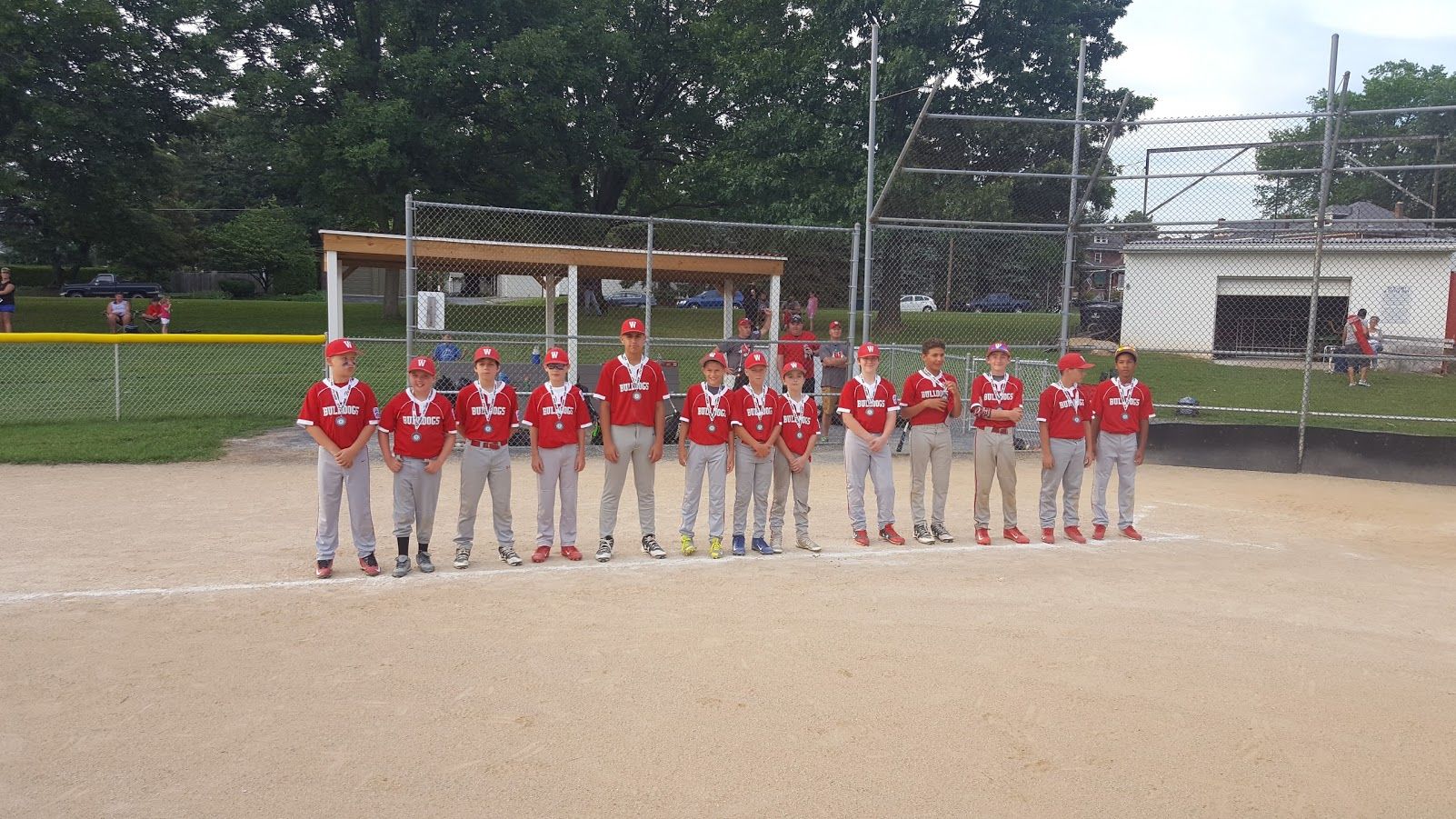 Youth baseball team in red jerseys lined up on a dirt field in front of a dugout.