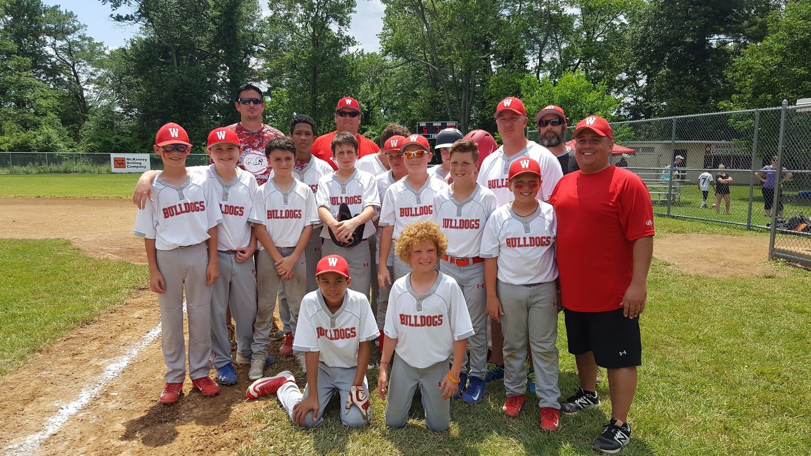 Youth baseball team posing on a field, wearing white uniforms and red caps with a coach in red.