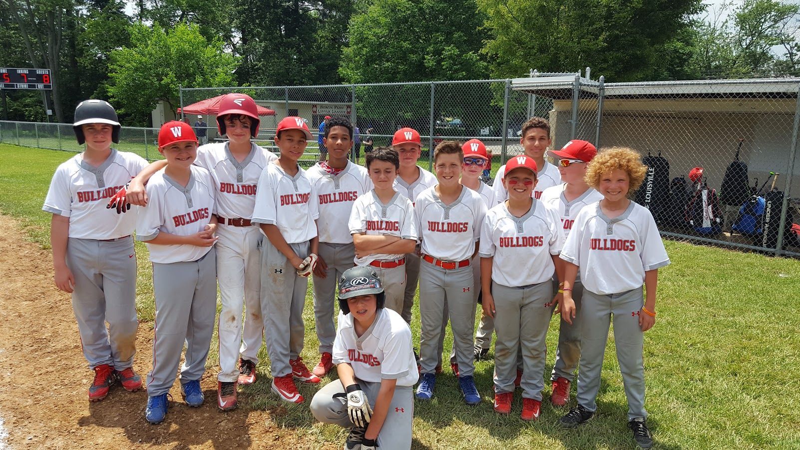 Youth baseball team posing on a field, wearing white uniforms and red caps, with trees and spectators in the background