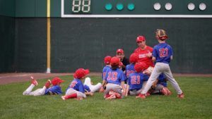 Youth baseball players in red and blue uniforms celebrate on the field after a play.