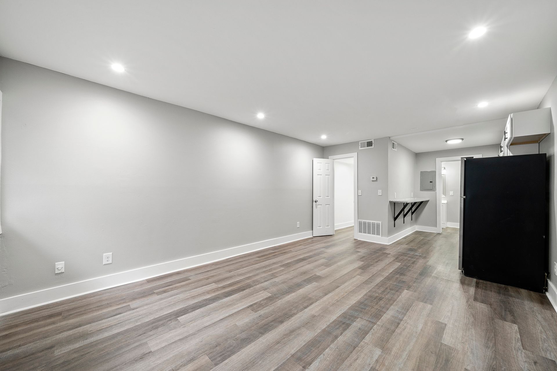 Empty modern living room with gray walls, wood-look floors, recessed lights, and a black appliance by the far wall