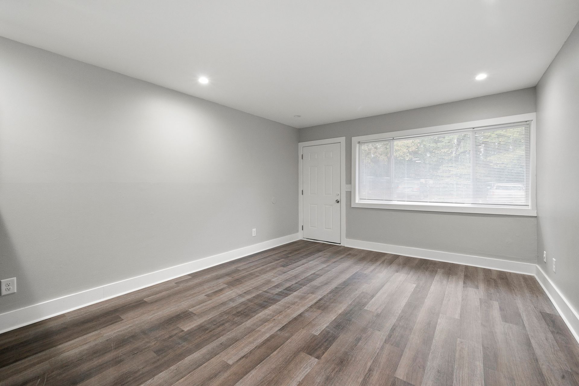 Empty gray room with wood flooring, recessed lights, a window, and a white door.