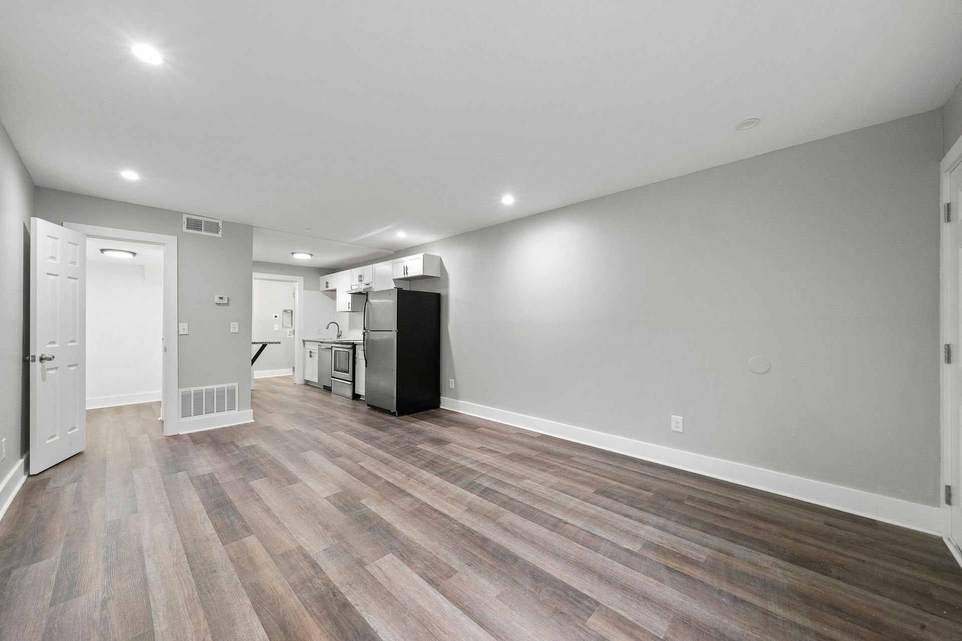 Empty modern living room with gray walls, wood-look floors, and an open kitchen in the background