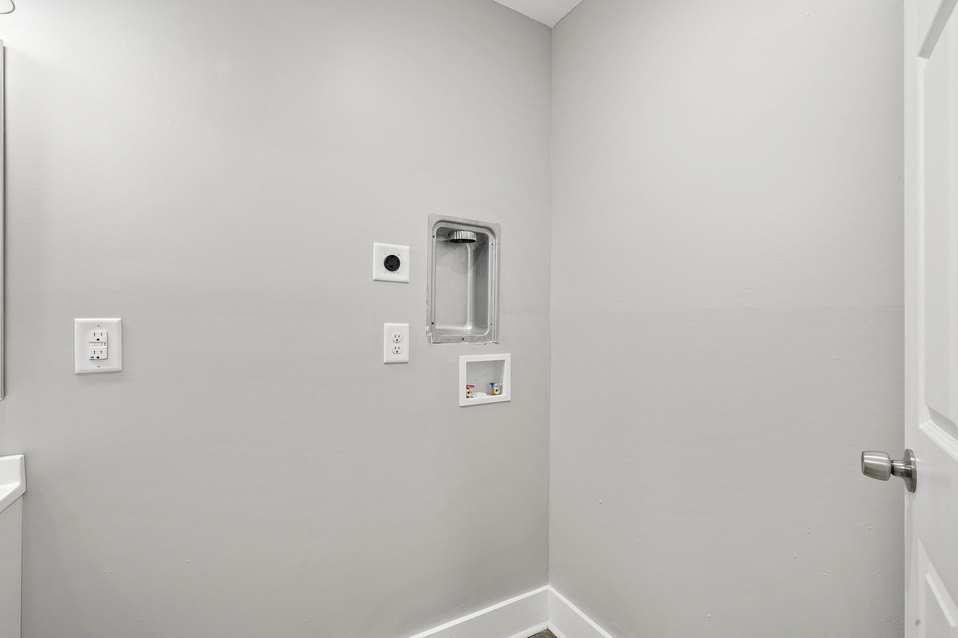 Empty laundry room wall with washer/dryer hookups, outlets, and a vent opening in light gray paint