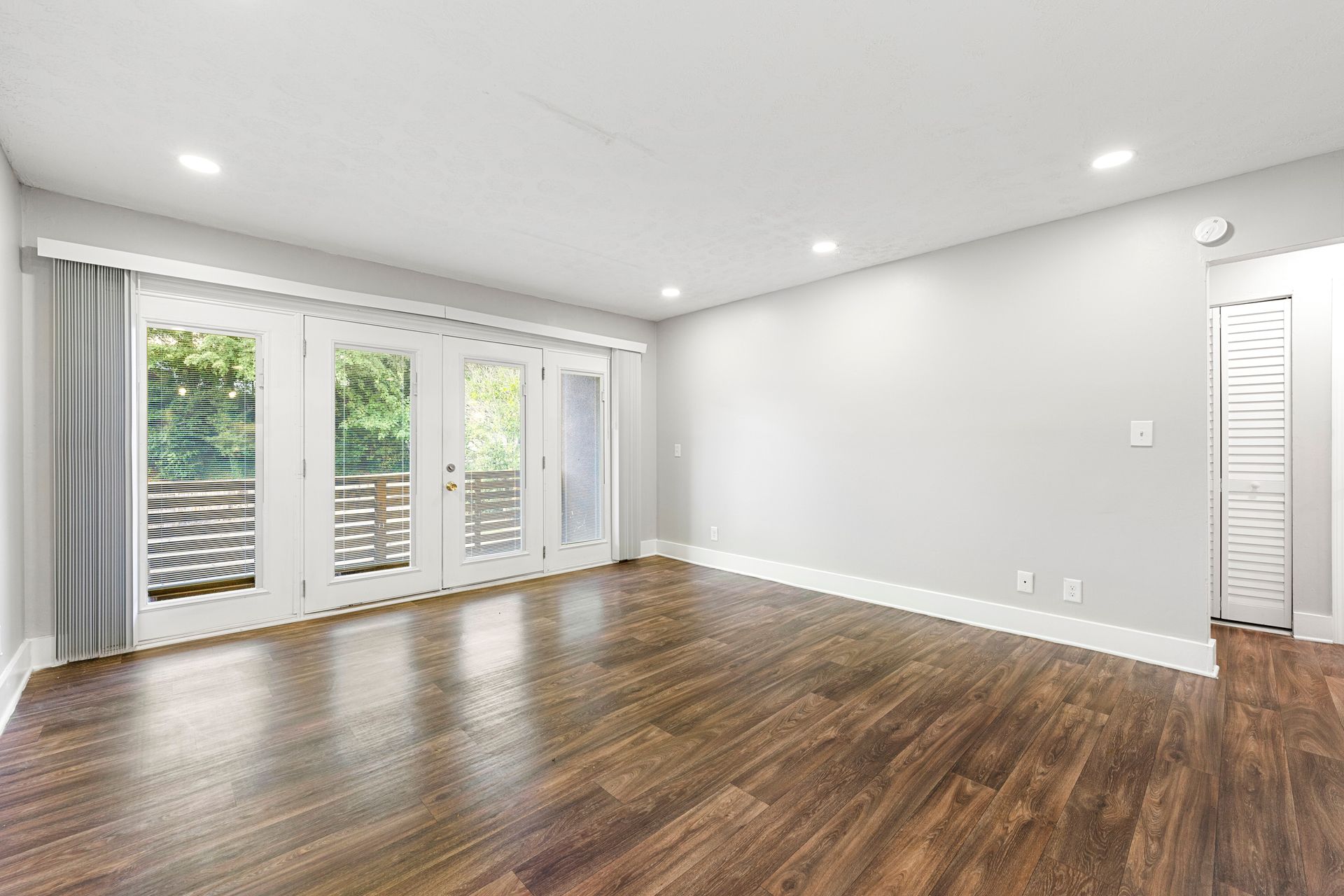Empty modern living room with hardwood floors, white walls, recessed lights, and glass doors to a balcony