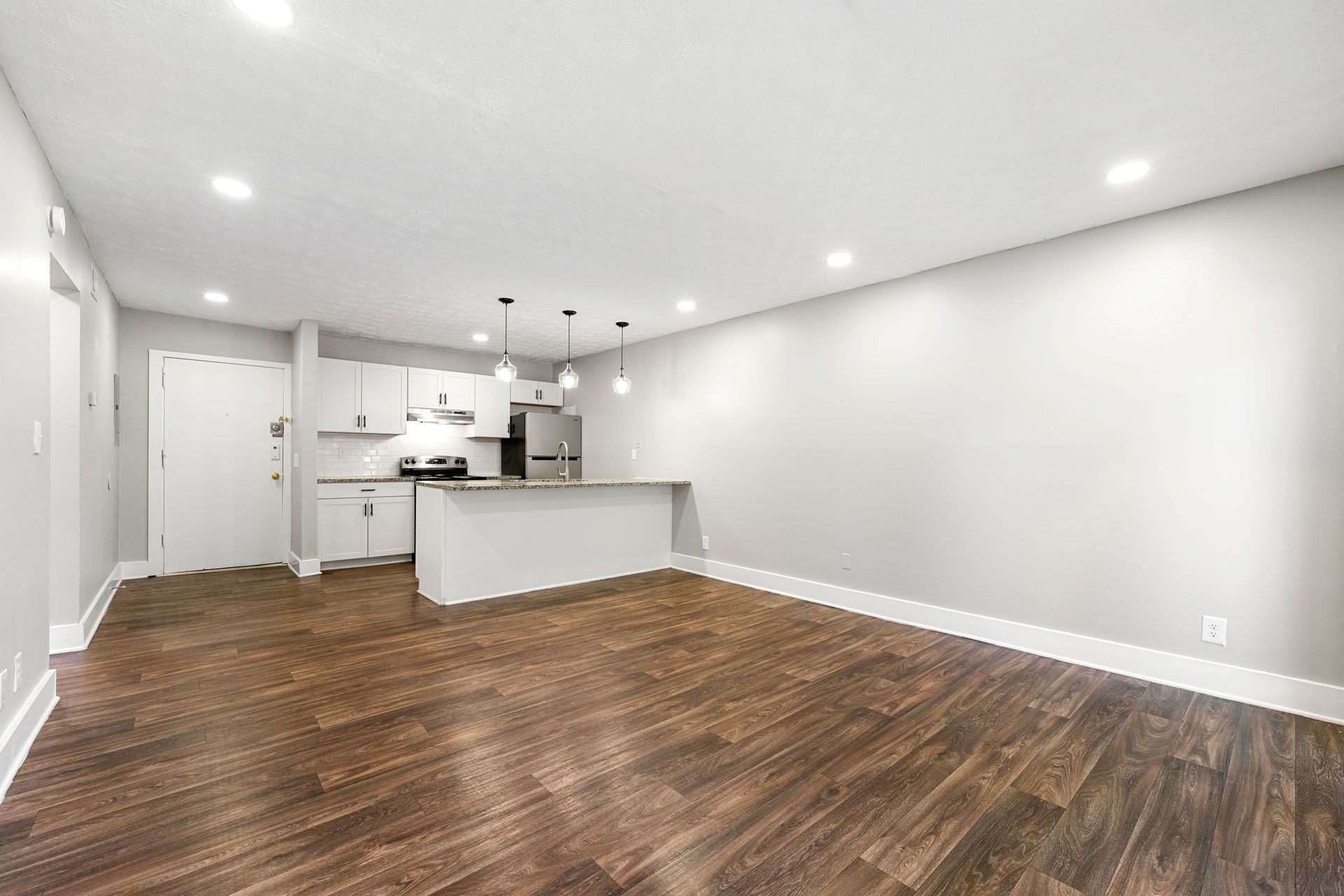 Spacious empty living room with dark wood floors, white walls, and a small open kitchen area.