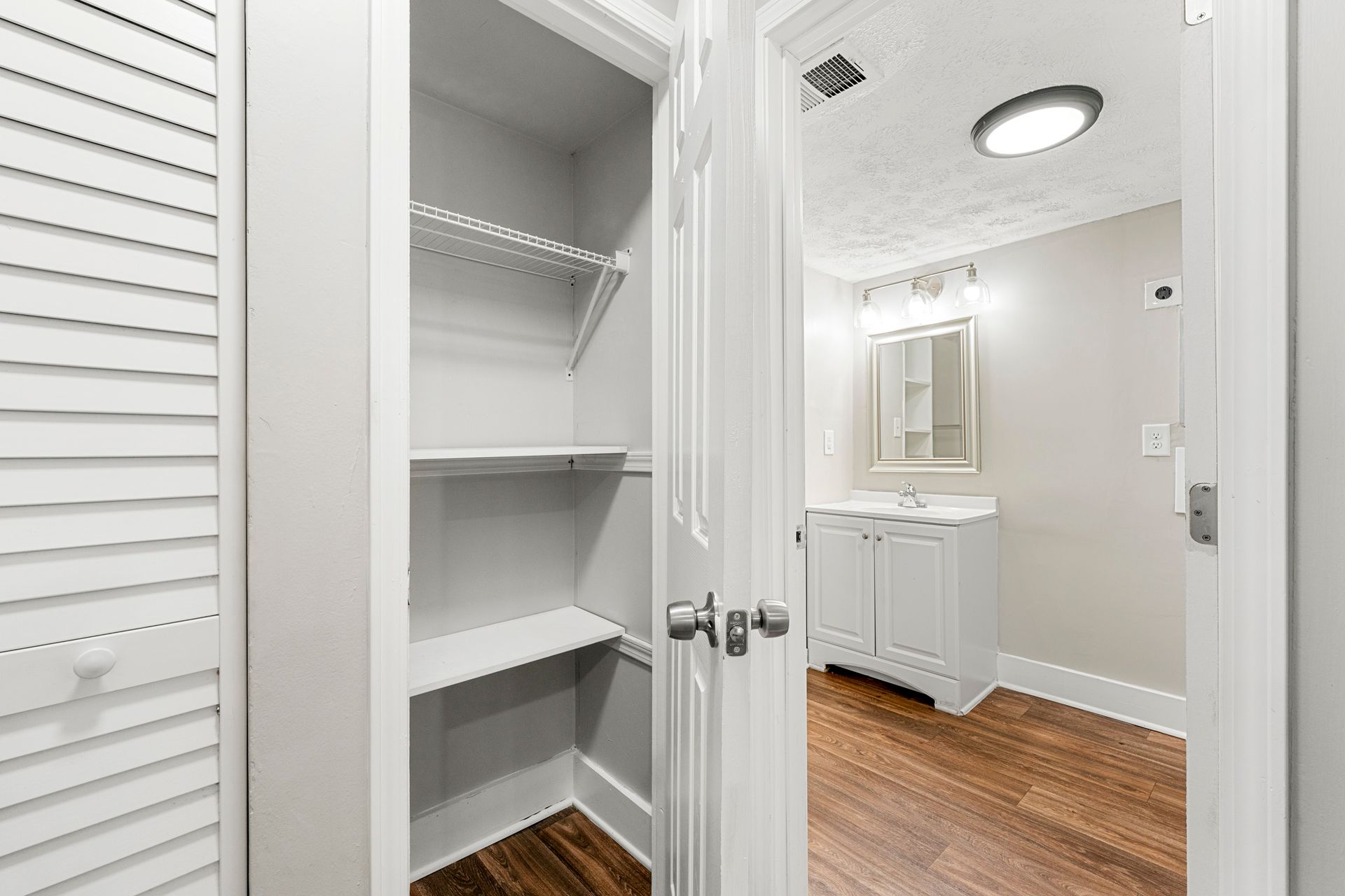 Open closet with white shelves beside a hallway vanity and mirror on hardwood floor