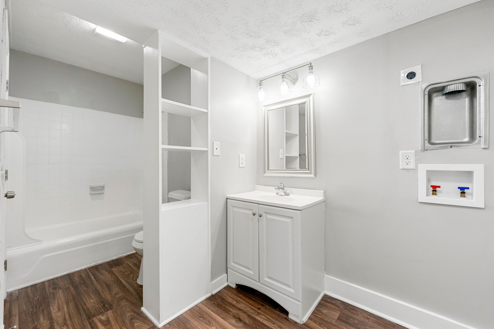 Bright bathroom with tub, white vanity and mirror, open shelving, and laundry hookups on the wall