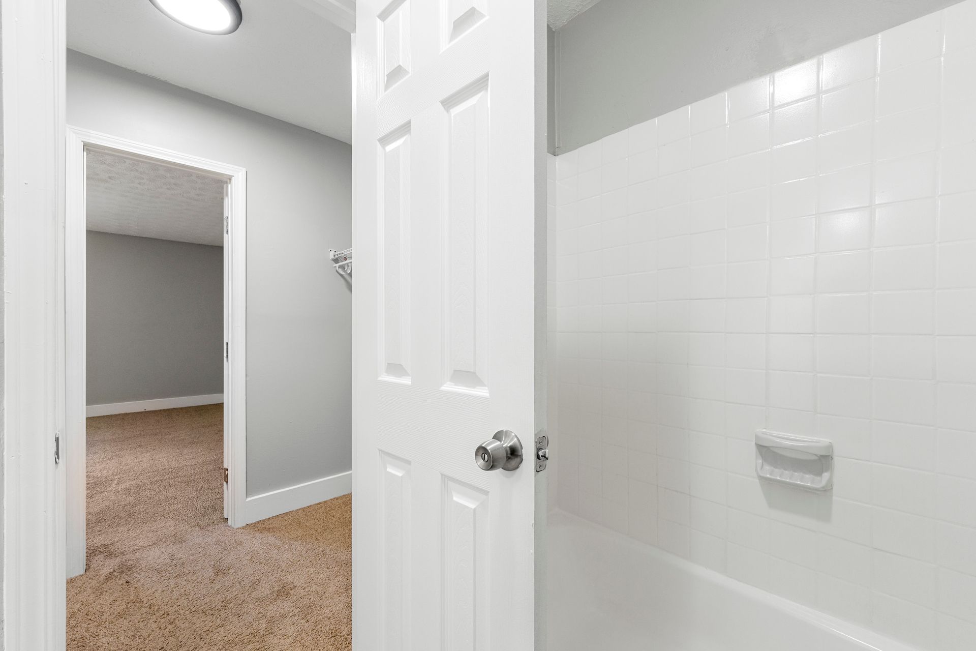 Bathroom doorway with white tiled shower wall, open door, and beige carpeted hall beyond
