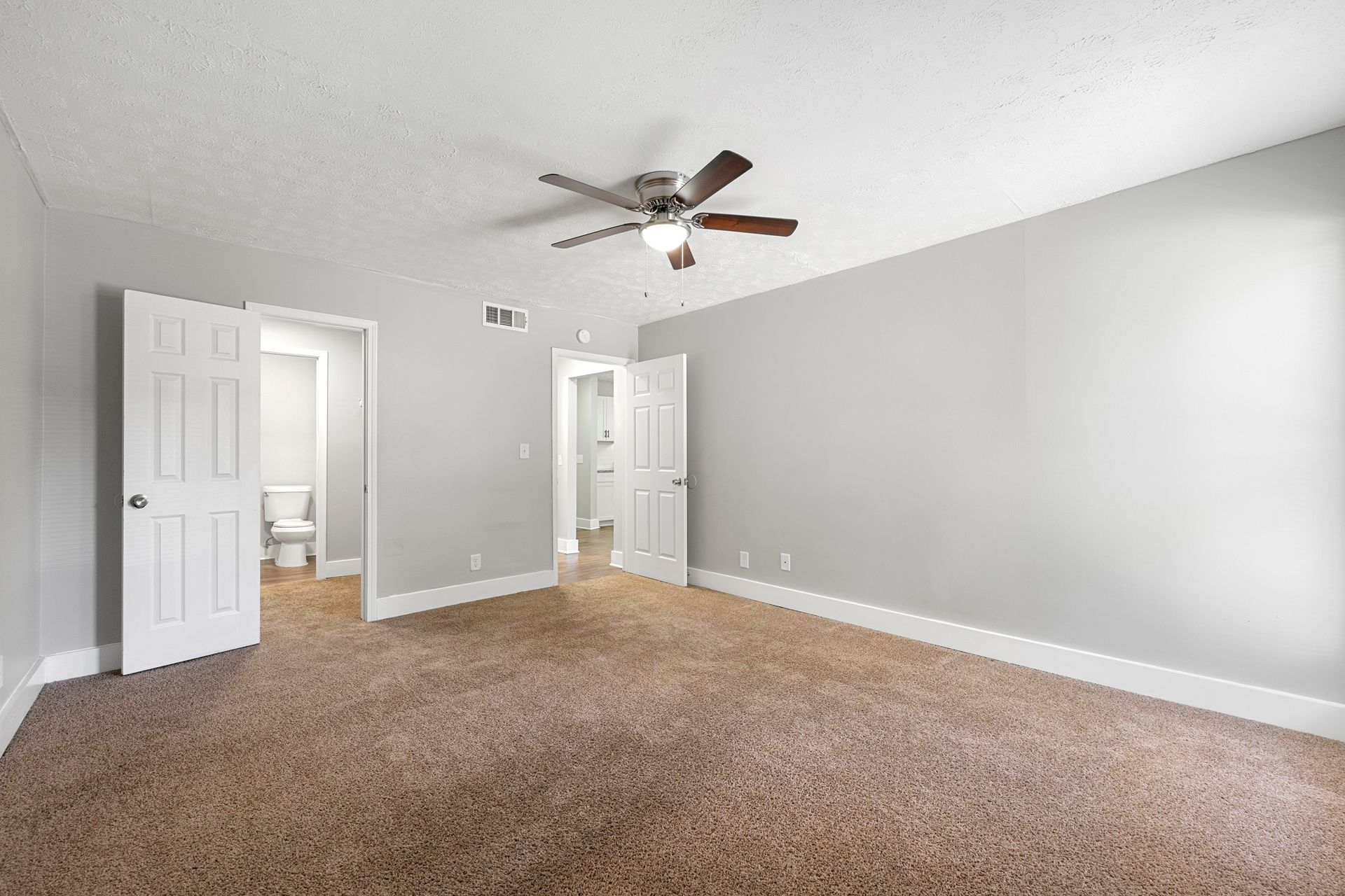 Empty gray bedroom with beige carpet, ceiling fan, and open doors to a hallway and bathroom.