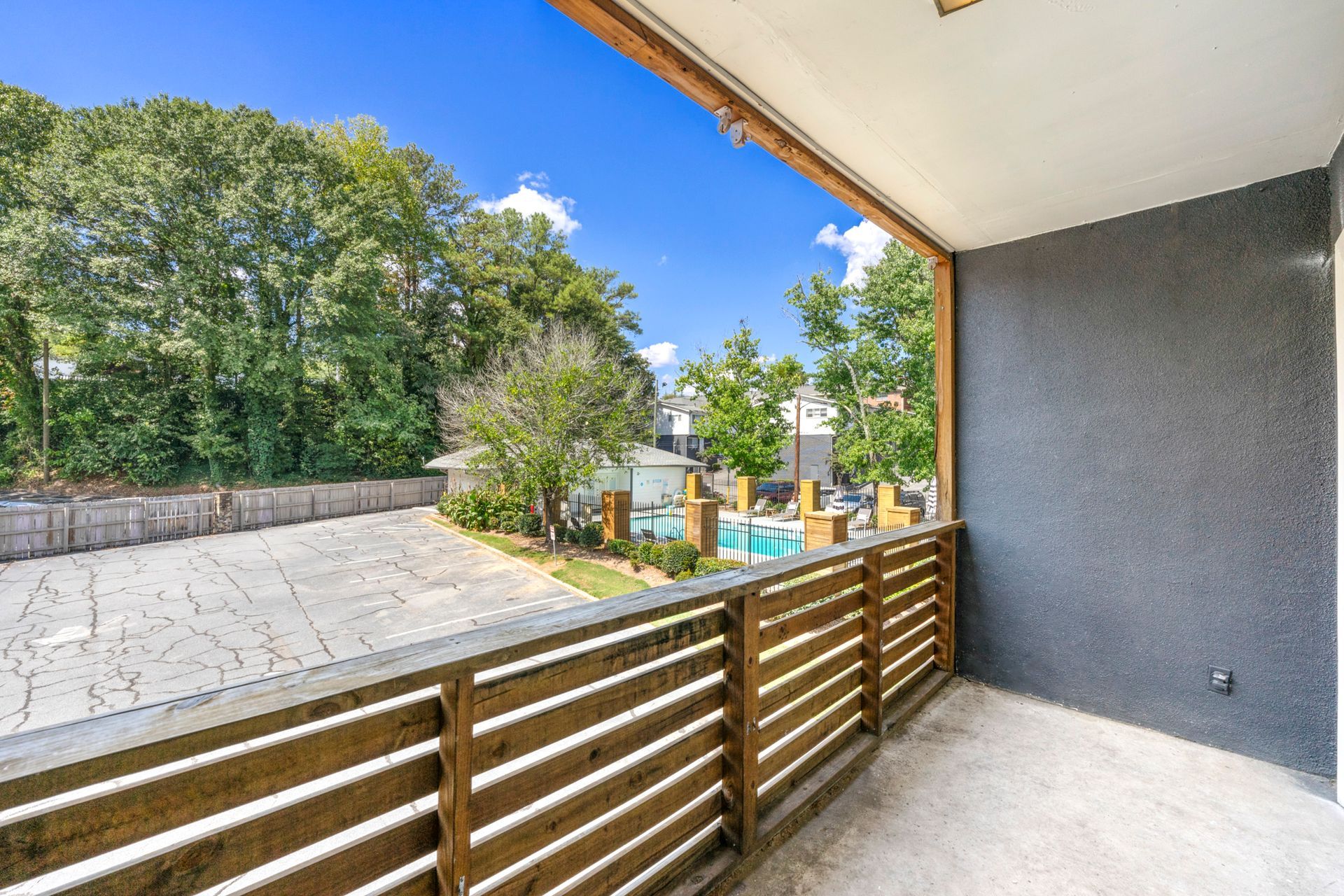 Balcony overlooking a paved courtyard, trees, and blue sky, with wooden railing and gray wall