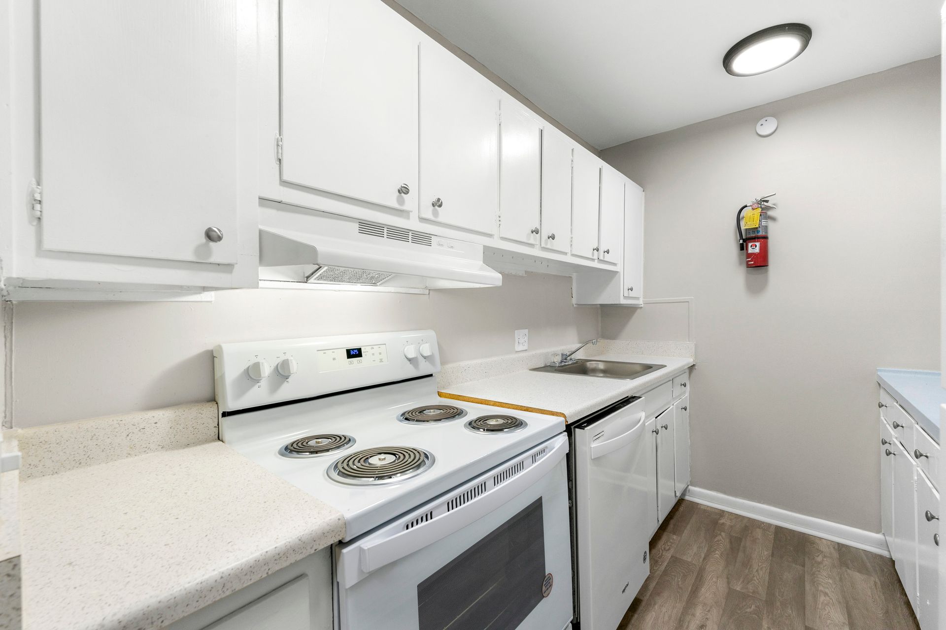 Bright white galley kitchen with electric stove, cabinets, countertop, and wood-look floor.