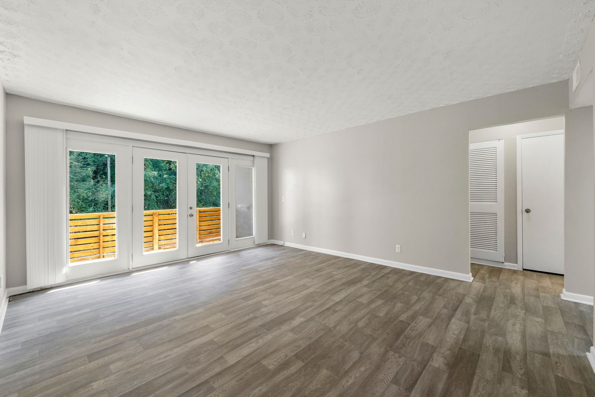 Empty modern living room with gray wood floors, white walls, and large glass doors to a deck.