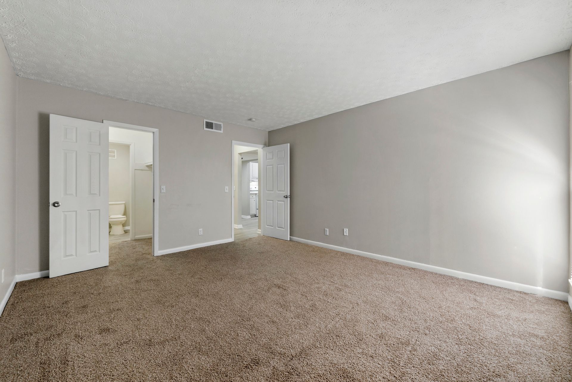 Empty beige bedroom with carpet, gray walls, and open white doors leading to a bathroom and closet.