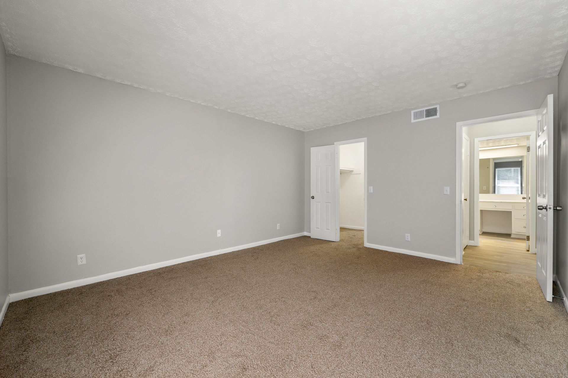 Empty beige-carpeted bedroom with gray walls and an open doorway to a hallway and adjoining room