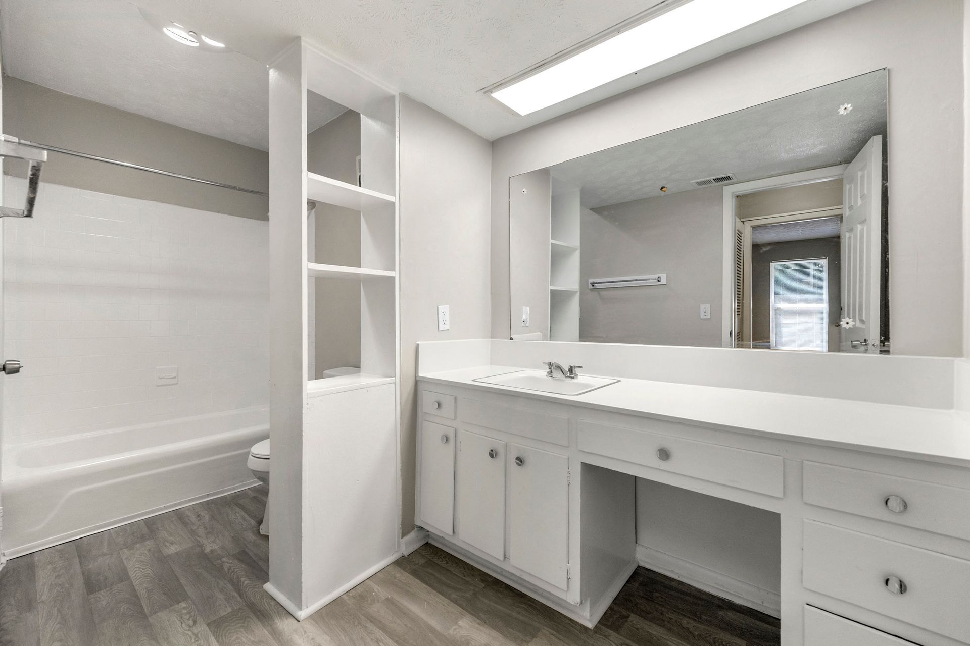 Bright modern bathroom vanity with large mirror, white cabinets, built-in shelves, and tub in the background