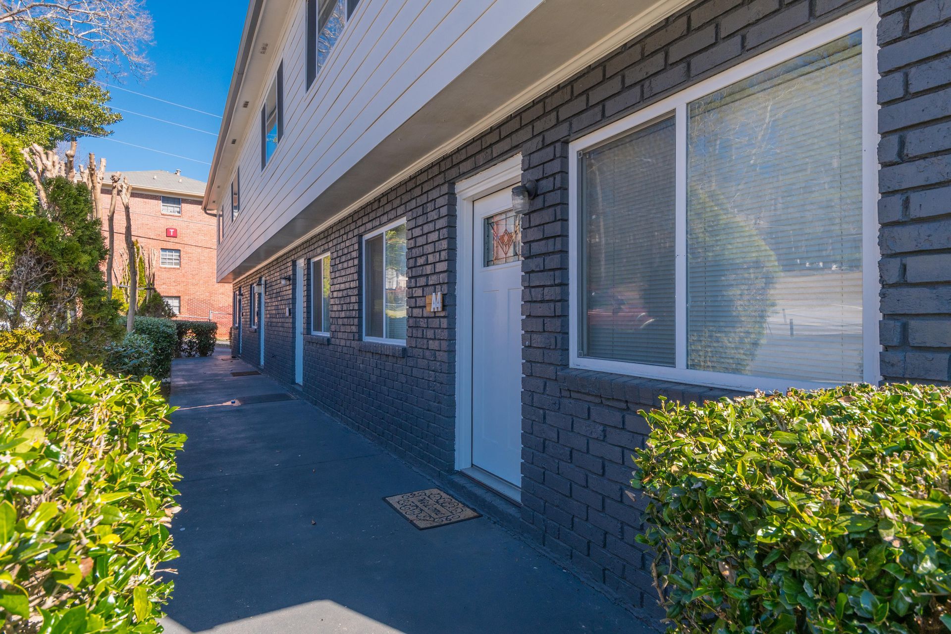 Apartment walkway with blue brick walls, white doors, windows, and trimmed hedges in bright sunlight