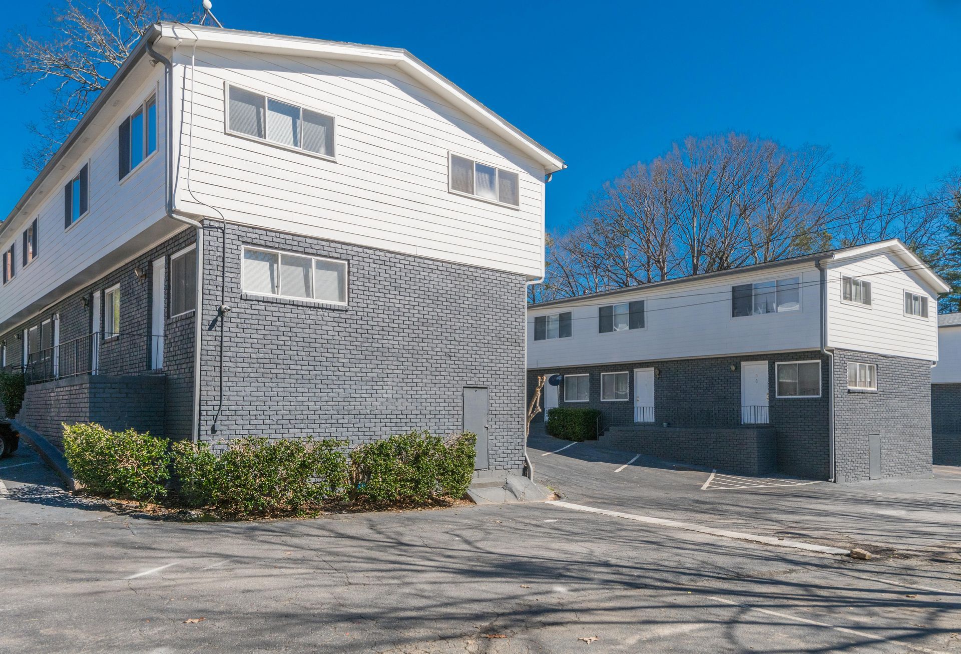 White multi-story apartment buildings with gray lower siding under a clear blue sky, viewed from a parking area