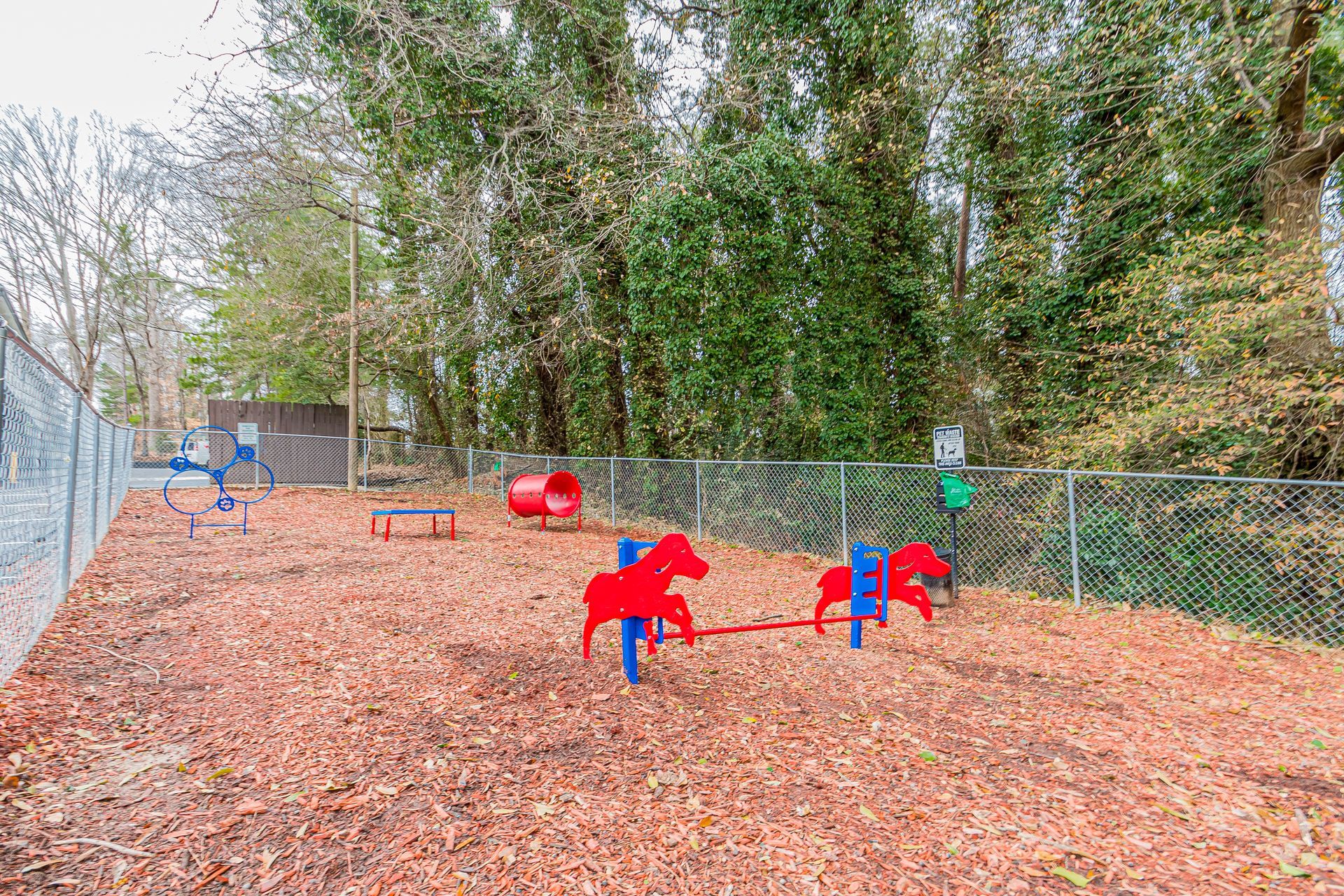 A fenced playground with red and blue play structures on a leaf-covered ground beside trees.