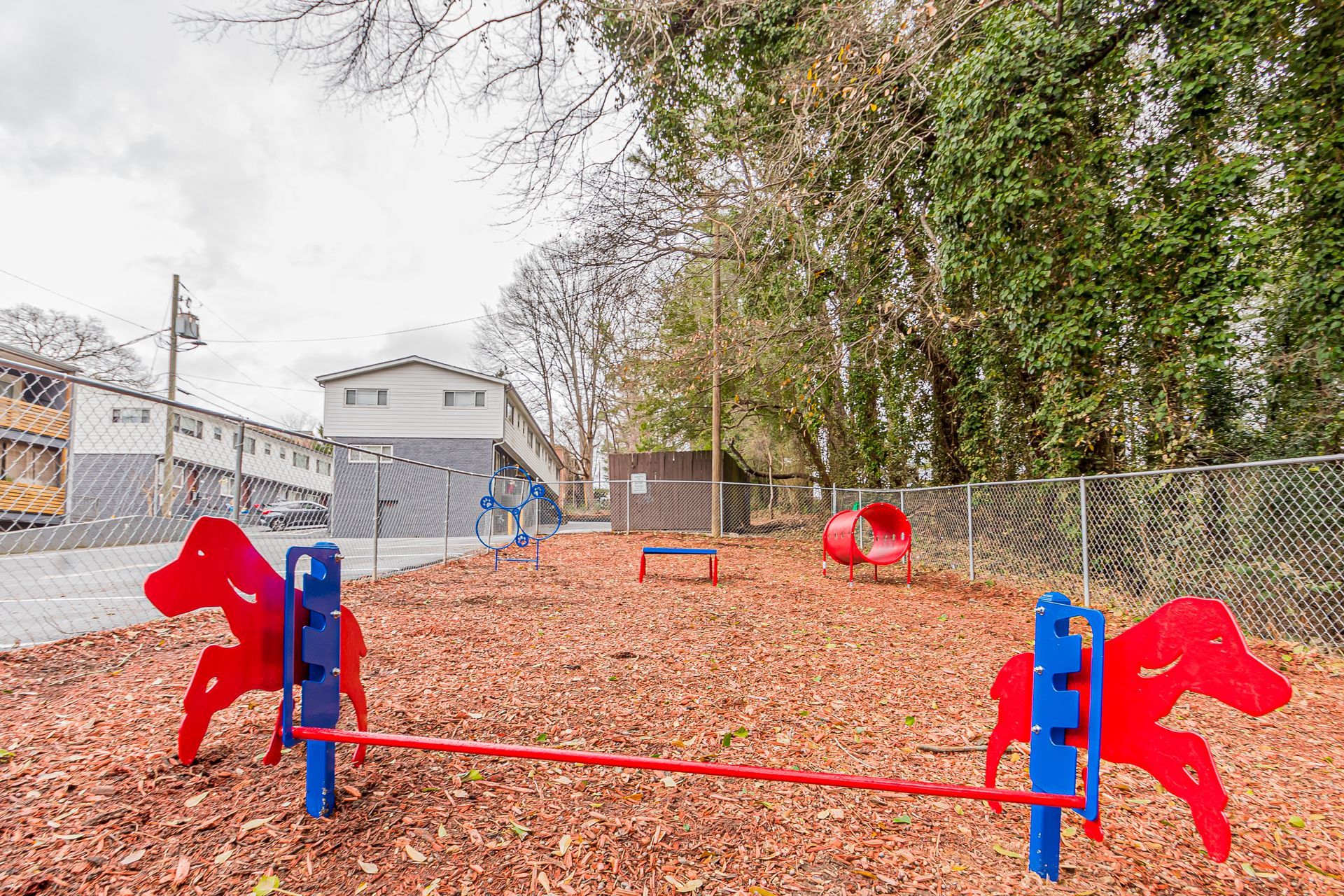Small outdoor playground with red and blue monkey bars on a leaf-covered ground near trees and buildings