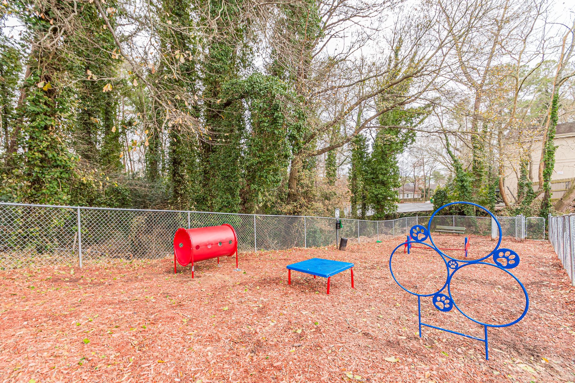 Small fenced playground with a red tunnel, blue platform, and blue climbing frame on wood chips.