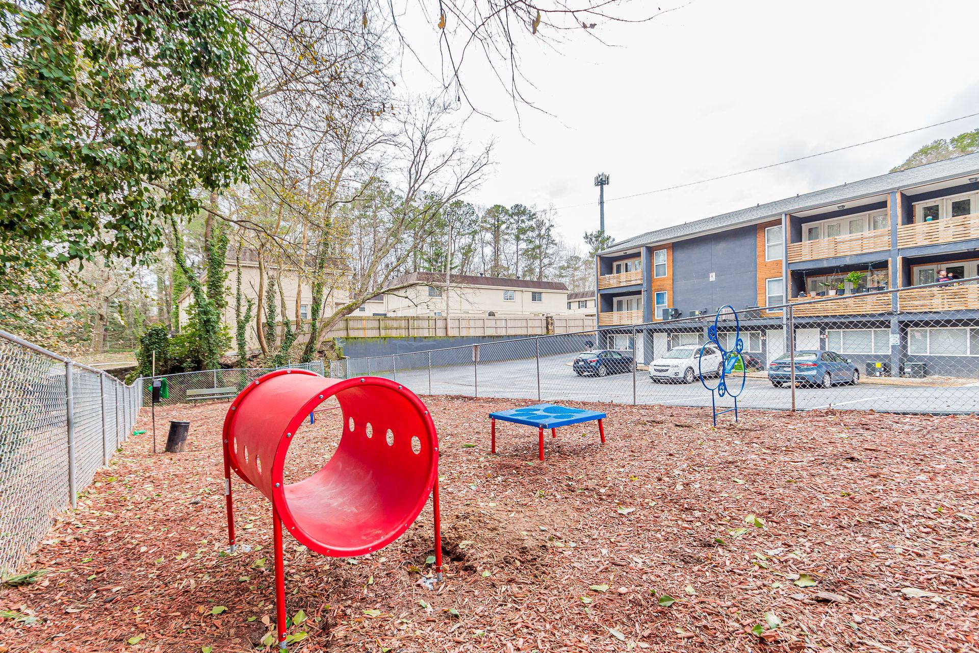 Red tube slide and blue seesaw in a leaf-covered playground beside apartment buildings