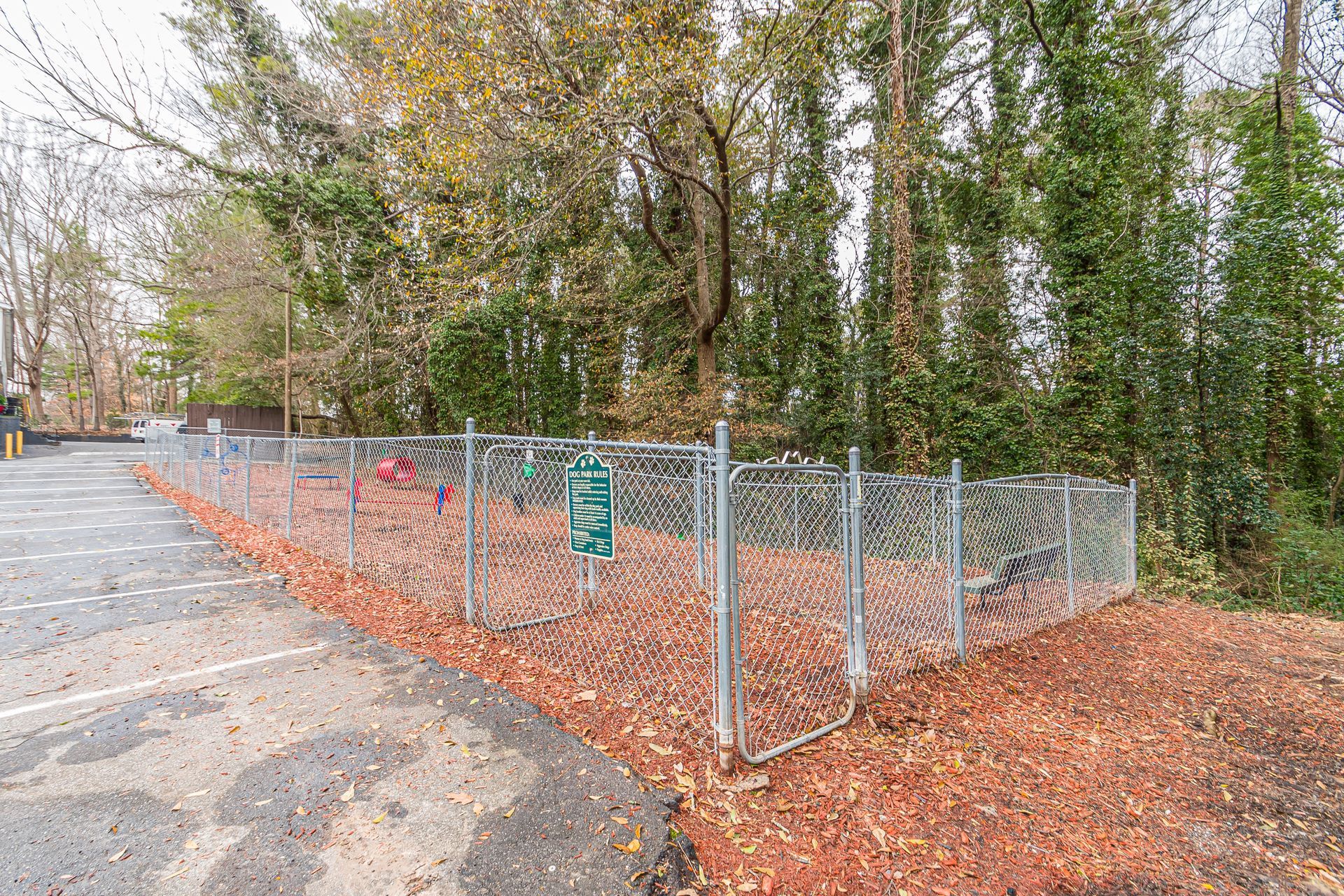 Chain-link fenced enclosure beside a wooded path, with orange leaf litter and a gate.