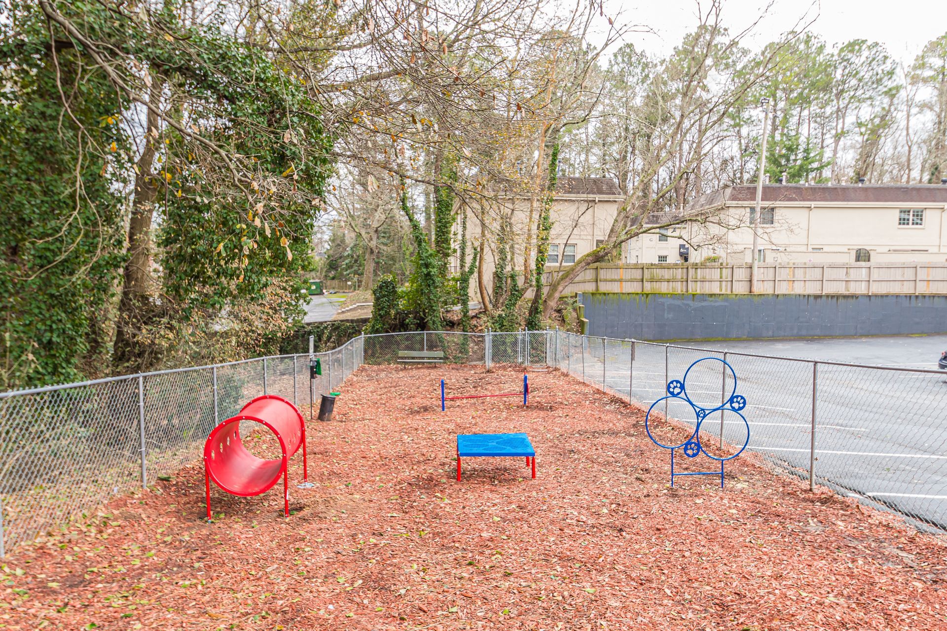Small playground with red tunnel, blue rocker, and climbing frame on wood chips beside a parking lot