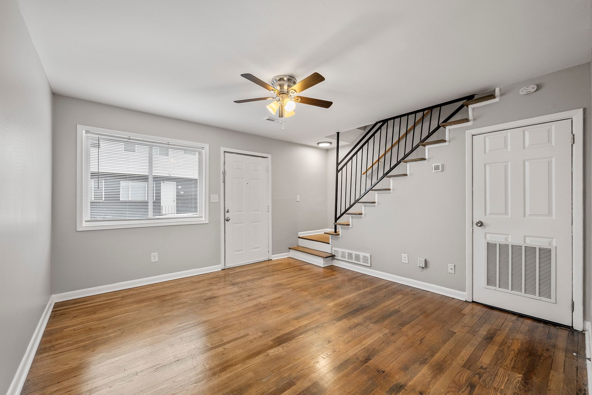 Empty living room with hardwood floors, ceiling fan, window blinds, and staircase railing in the corner