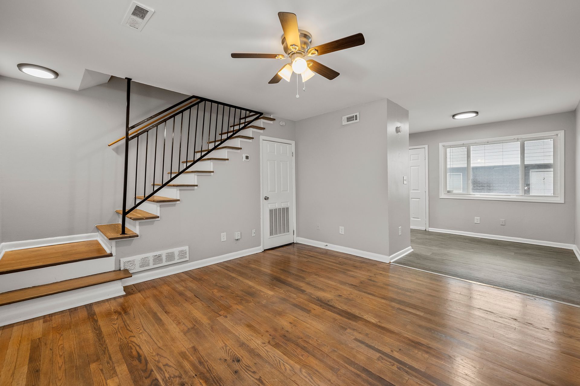 Empty living room with hardwood floors, staircase, ceiling fan, and a carpeted alcove by a window.
