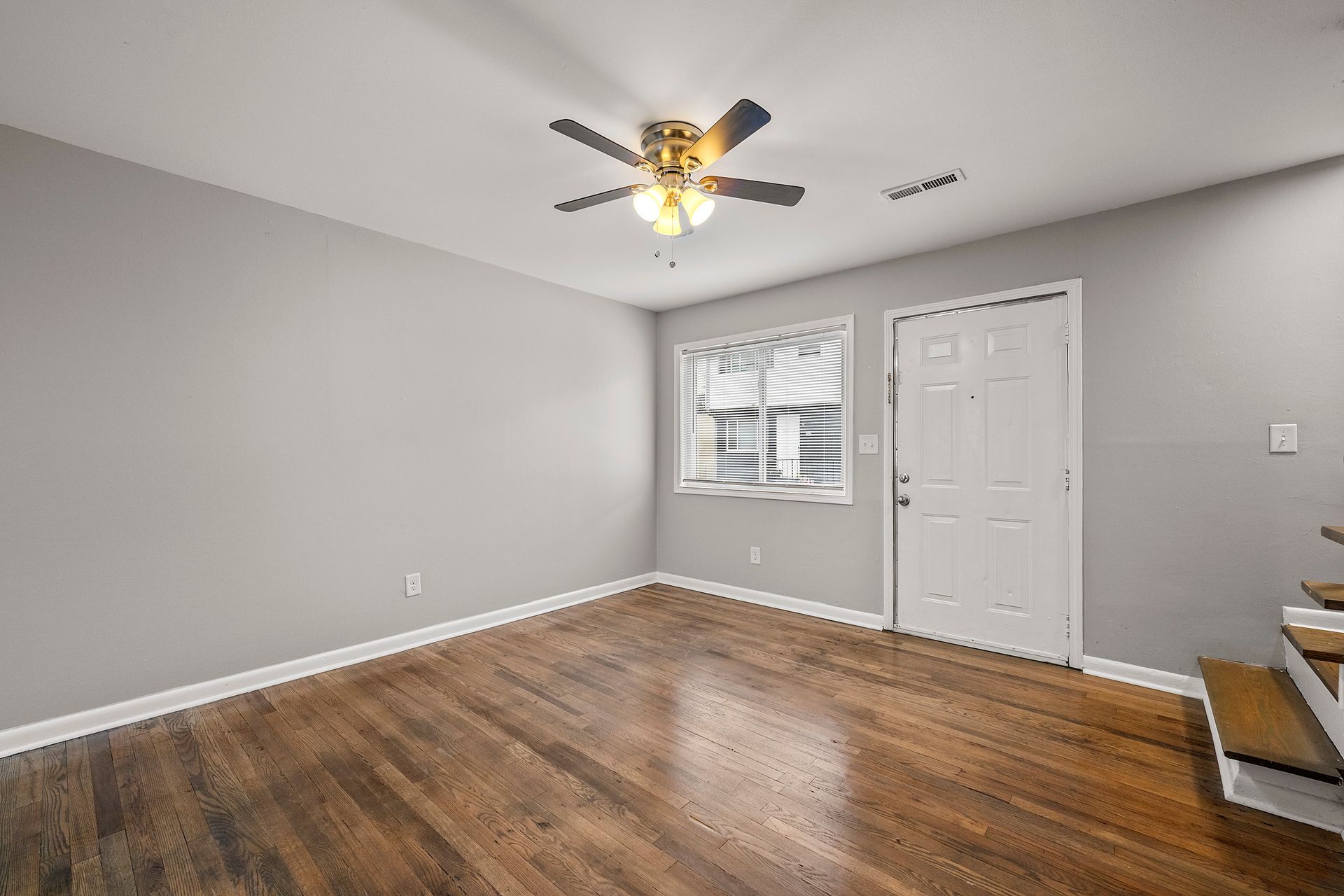 Empty gray bedroom with hardwood floors, ceiling fan, window, white door, and staircase at right