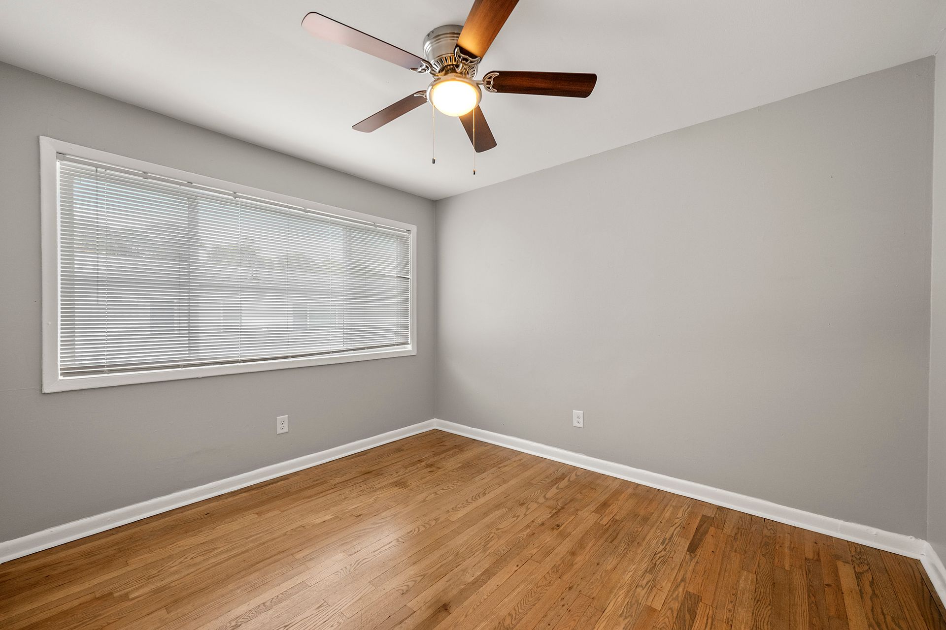 Empty gray bedroom with hardwood floor, window blinds, and a ceiling fan with light.