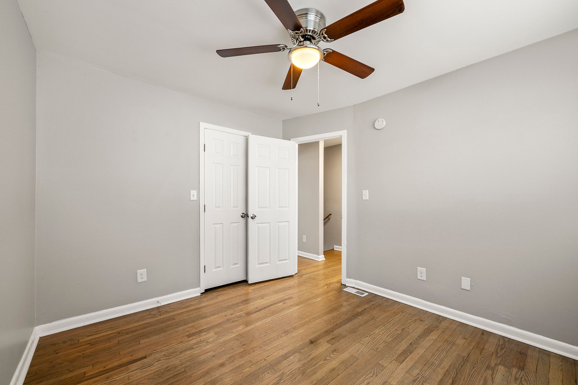 Empty bedroom with gray walls, wood floors, white closet doors, and a ceiling fan.