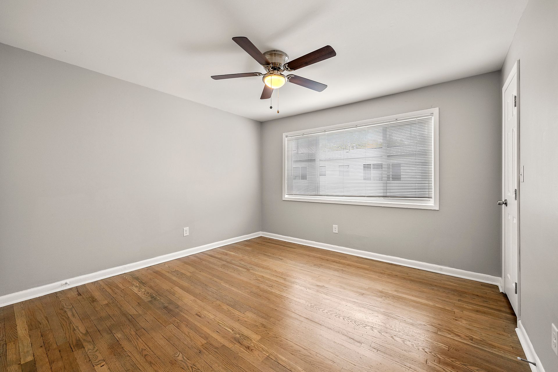 Empty room with gray walls, hardwood floor, ceiling fan, and a window with blinds.