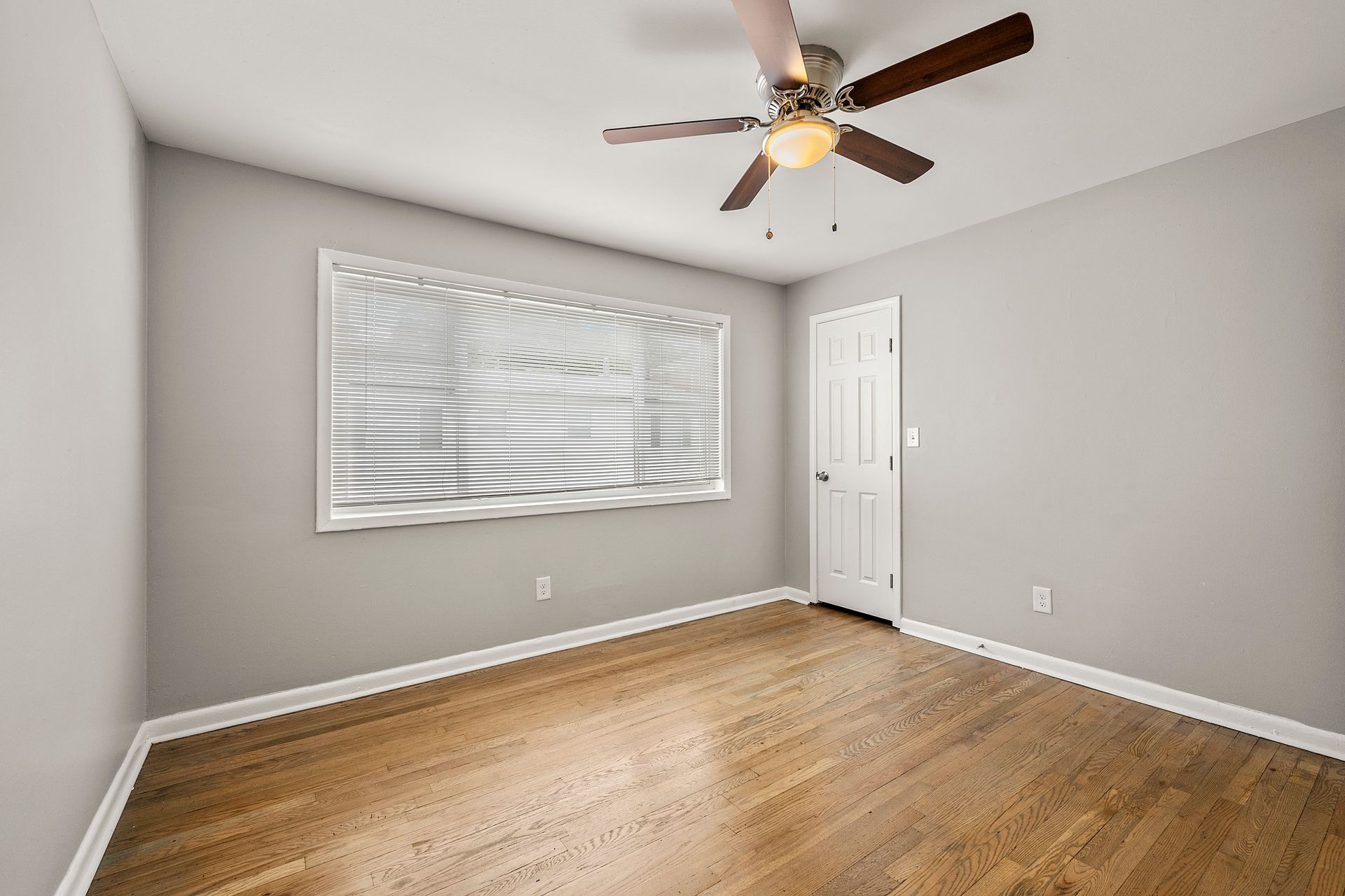 Empty gray bedroom with hardwood floor, window blinds, white door, and ceiling fan