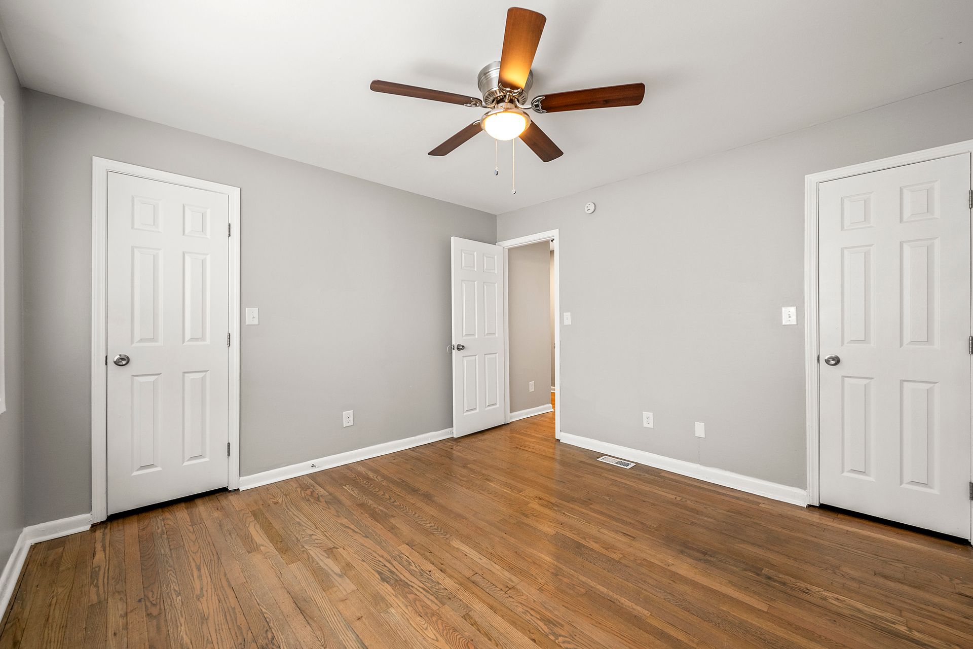 Empty gray bedroom with hardwood floors, white closet doors, and a ceiling fan with light.