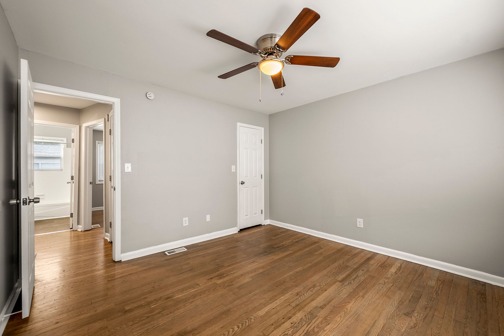 Empty bedroom with gray walls, wood floors, ceiling fan, and open doorway to a hallway