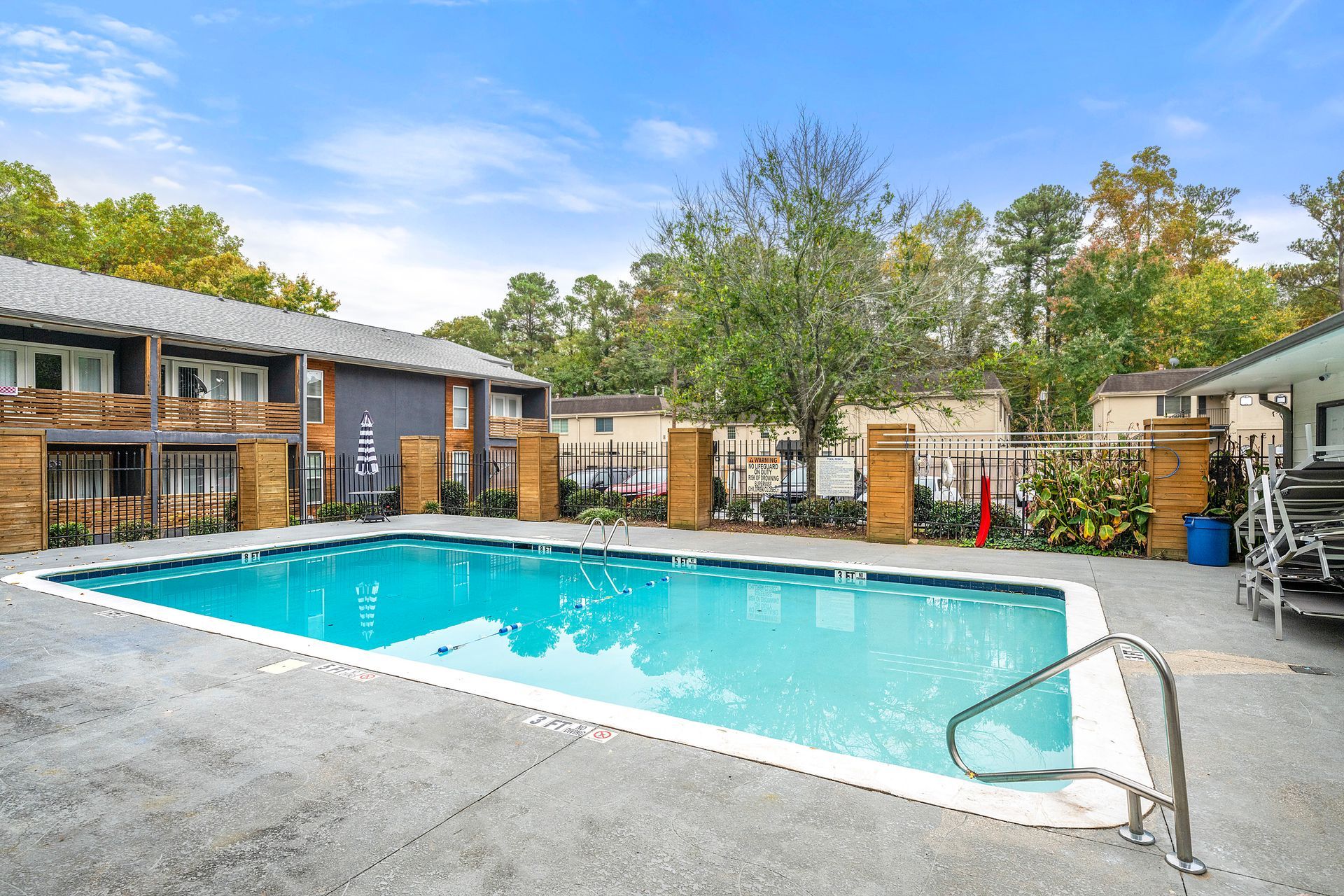 Outdoor apartment pool with turquoise water, concrete deck, lounge chairs, and surrounding trees under a blue sky