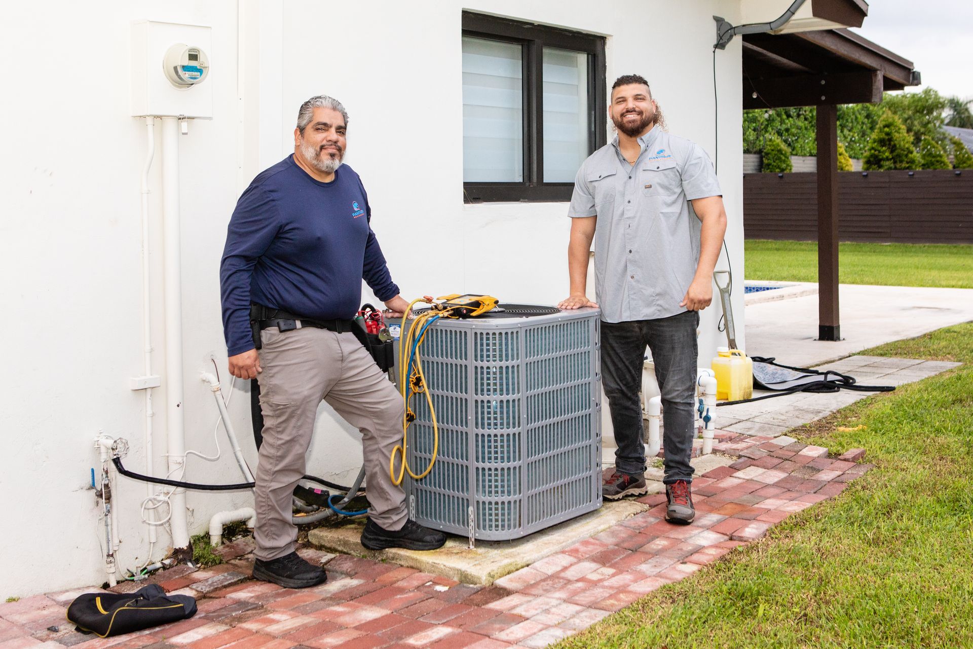 Two men stand beside an outdoor HVAC unit beside a white house and brick patio.