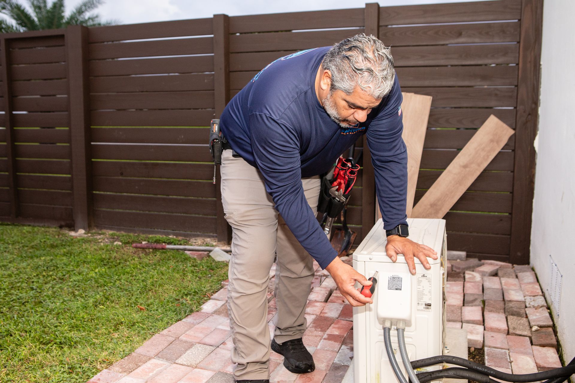 Person adjusting a white outdoor appliance on a patio beside a wooden fence