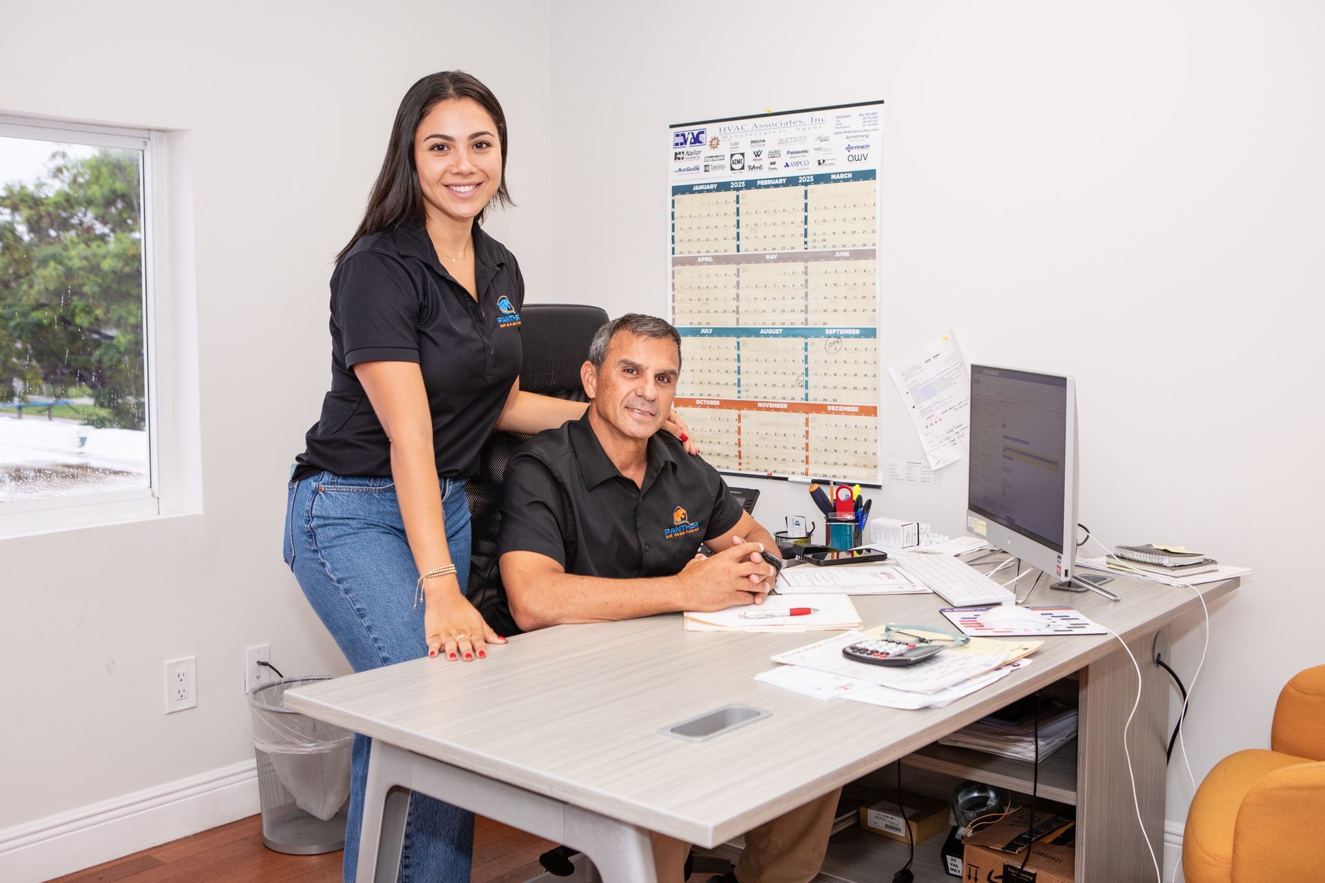 Two coworkers smiling in a small office, standing and sitting beside a desk with a computer and calendar.
