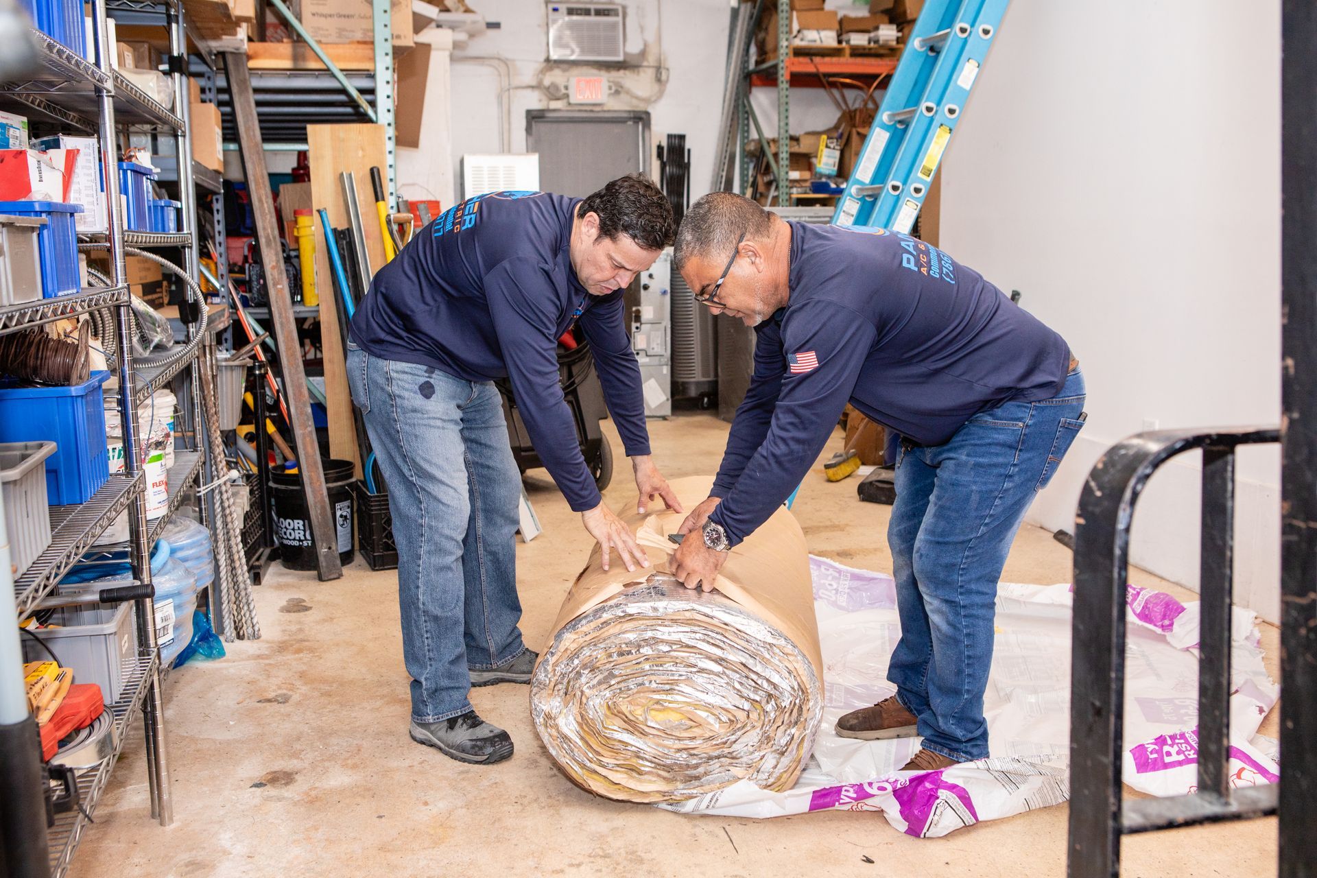 Two workers in a workshop open a large wrapped package on the floor.
