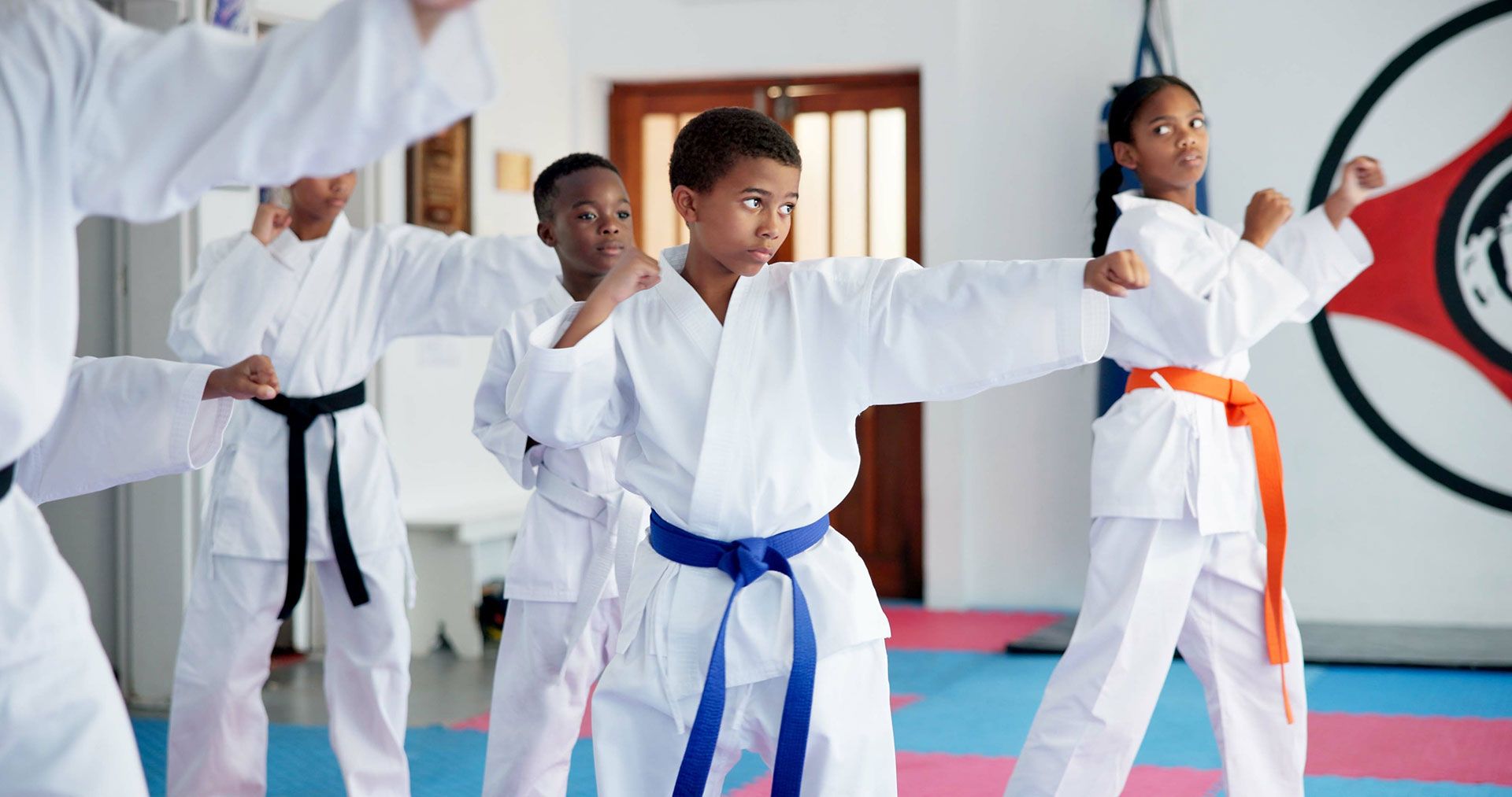 Children in white karate uniforms practicing martial arts in a dojo, one with a blue belt and others behind her